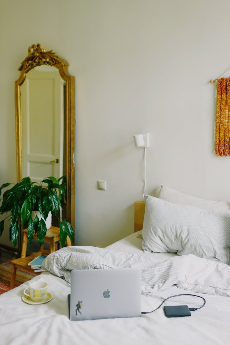 High angle of modern opened laptop with power bank and cup of hot drink placed on comfortable bed with soft blanket and pillows in stylish bedroom in daylight